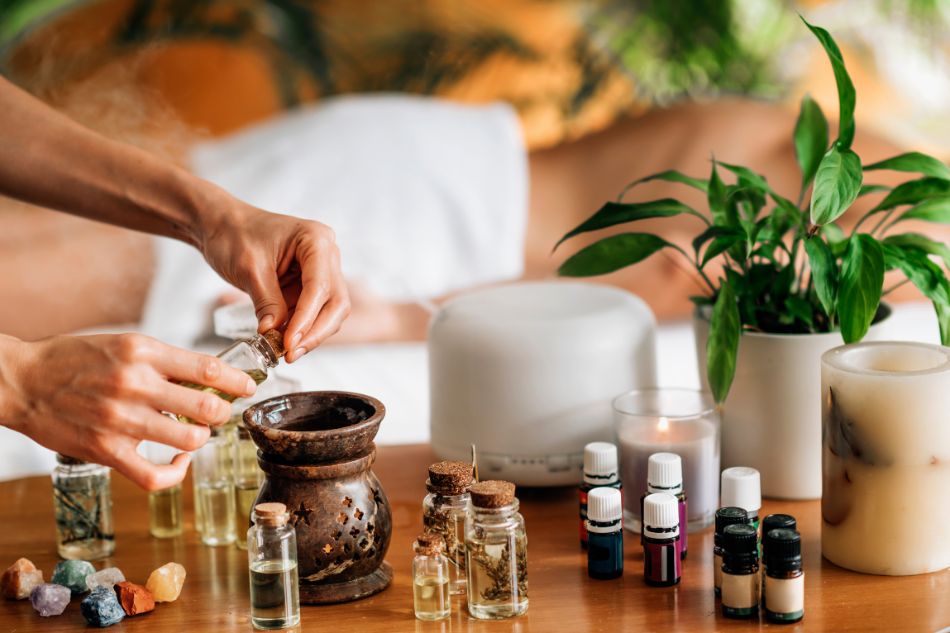 Pessoa adicionando óleo essencial a um difusor, com óleos, cristais e vela ao fundo em um ambiente de spa.