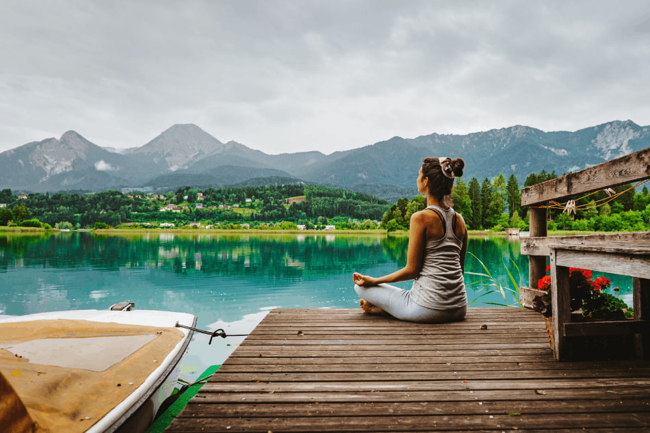 Pessoa meditando em um deck de madeira à beira de um lago, com montanhas ao fundo em um dia nublado.
