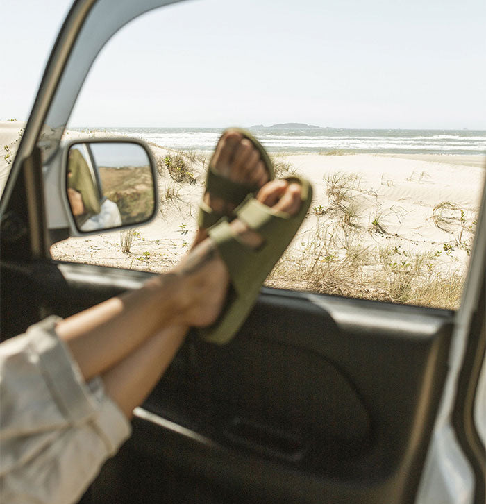 Pernas cruzadas com sandália na cor verde apoiadas sobre a porta do carro de frente para o mar.