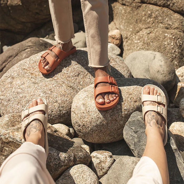 Duas pessoas com os pés em cima de rochas usando sandálias na cor laranja e bege.
