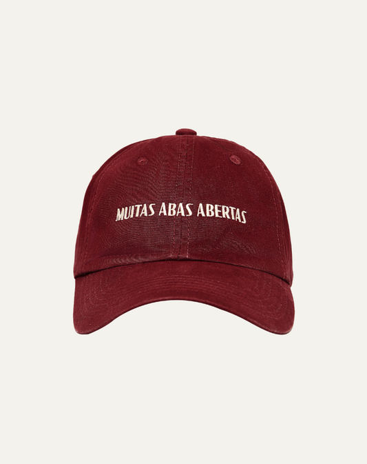 Boné em algodão na cor vermelho bordô com bordado escrito muitas abas abertas em off white, vista frontal no fundo cinza claro.