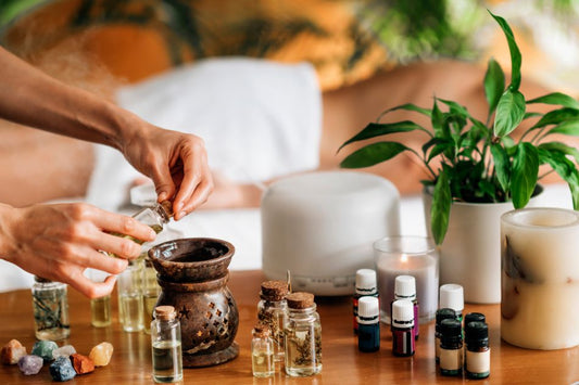 Pessoa adicionando óleo essencial a um difusor, com óleos, cristais e vela ao fundo em um ambiente de spa.