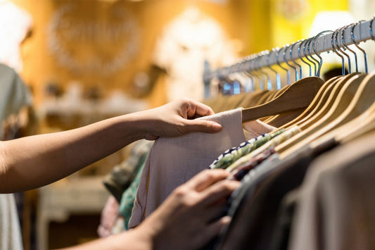Pessoa pegando uma peça de roupa de uma arara em uma loja de roupas, com várias opções de roupas ao fundo.