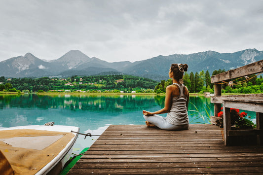 Pessoa meditando em um deck de madeira à beira de um lago, com montanhas ao fundo em um dia nublado.