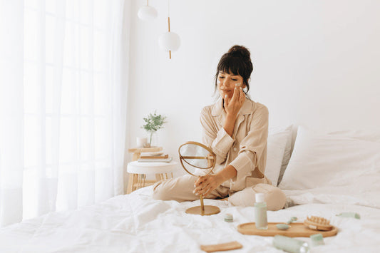 Mulher de pijama aplicando creme no rosto em um espelho de mão, sentada na cama com produtos de cuidados com a pele ao seu redor.