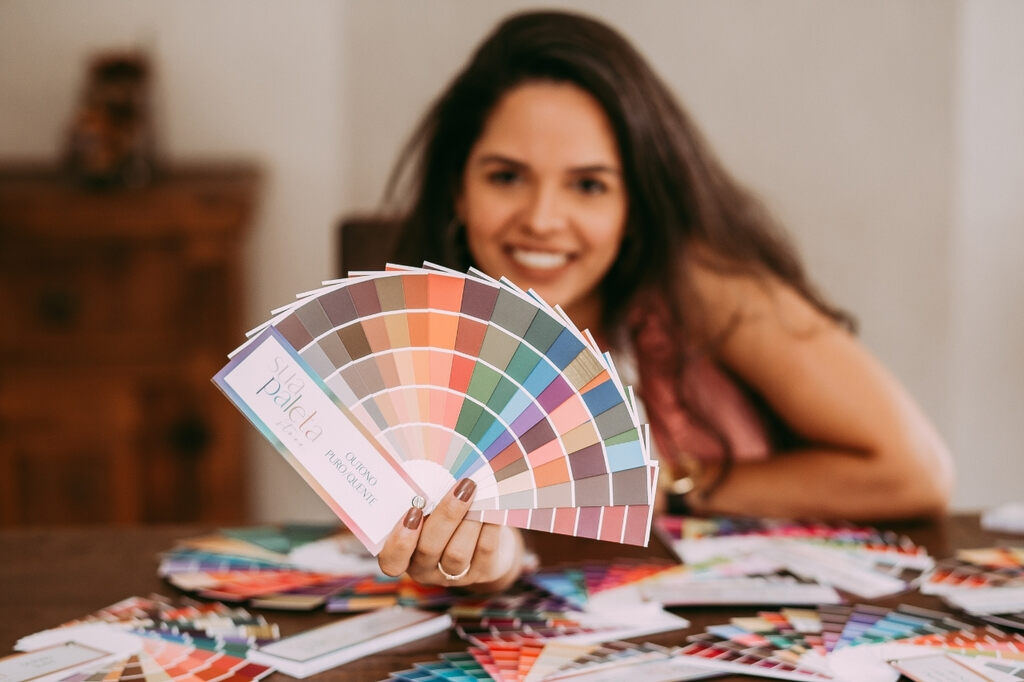Mulher segurando cartela de cores com diferentes tonalidades e várias paletas de cores espalhadas sobre a mesa para ilustrar a coloração pessoal e as possíveis combinações 