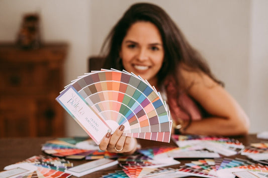 Mulher segurando cartela de cores com diferentes tonalidades e várias paletas de cores espalhadas sobre a mesa para ilustrar a coloração pessoal e as possíveis combinações 