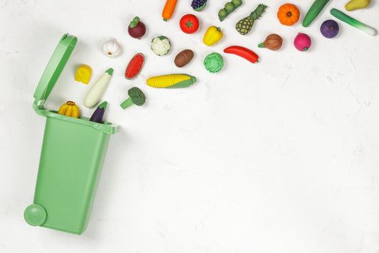 Imagem de uma lixeira verde aberta com diversos legumes e frutas coloridos saindo dela sobre um fundo branco, representando descarte de alimentos.