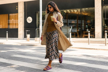 Mulher usando um dos clássicos looks de inverno, com sobretudo bege e saia xadrez em passeio na rua