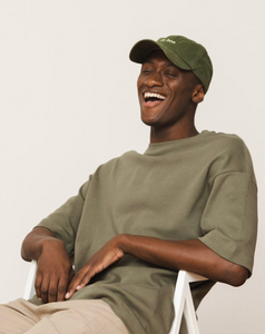 Homem sorrindo enquanto usa um boné verde com a frase "de boa" bordada na frente. Ele veste uma camiseta larga verde oliva e está sentado de forma relaxada em uma cadeira.