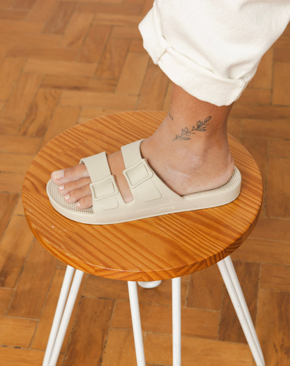 Pessoa em pé sobre um banquinho de madeira, vestindo as sandálias bege claro. O foco está nos pés e no design das sandálias, com uma tatuagem visível no tornozelo.