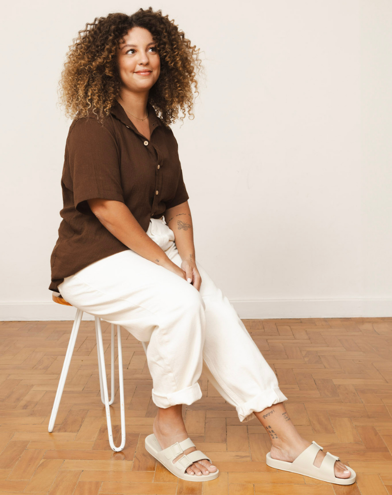 Mulher sentada em uma cadeira, vestindo uma camisa marrom e calça branca, combinada com as sandálias bege claro. Ela parece relaxada, olhando para o lado com um leve sorriso.