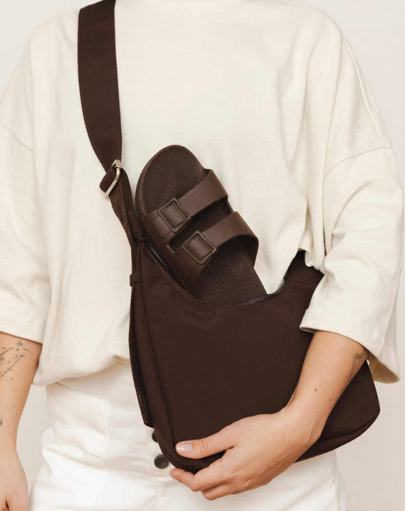 Pessoa carregando uma bolsa marrom com uma sandália combinando guardada em um dos compartimentos. 