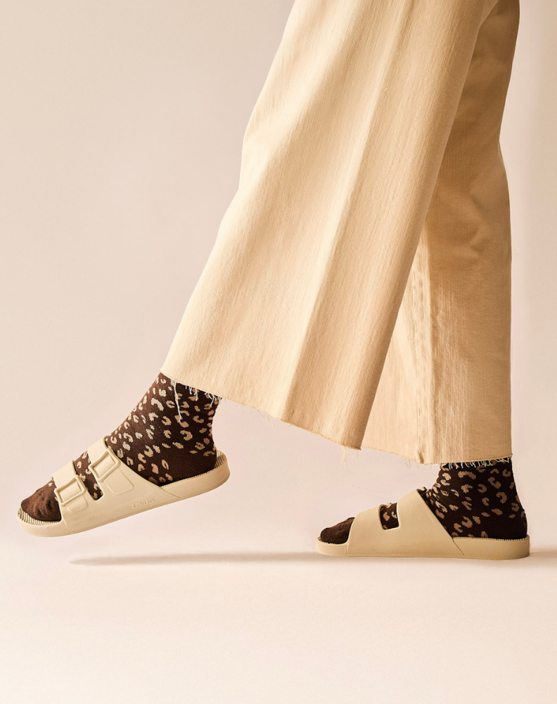 Pessoa usando o par de meias de algodão marrom escuro café com estampa de onça marrom clara e sandália Linus off white baunilha, com o pé direito apoiado no chão e o esquerdo levantado, calça bege clara de barra larga aparente até o joelho. Fundo da imagem rosado. 