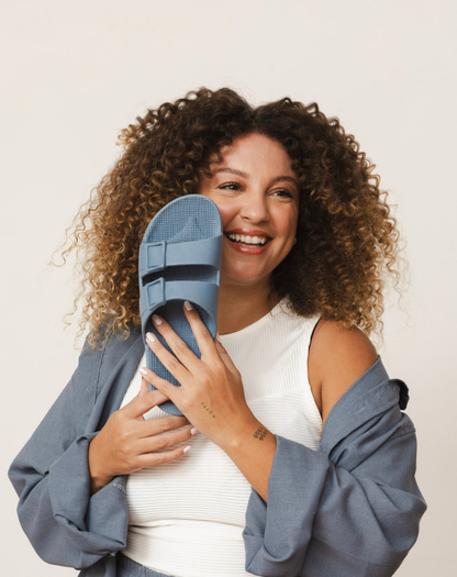 Mulher sorridente, com cabelos cacheados volumosos, segura uma sandália azul próxima ao rosto. Ela está vestindo um look casual e despojado, com uma blusa branca e um casaco azul aberto.