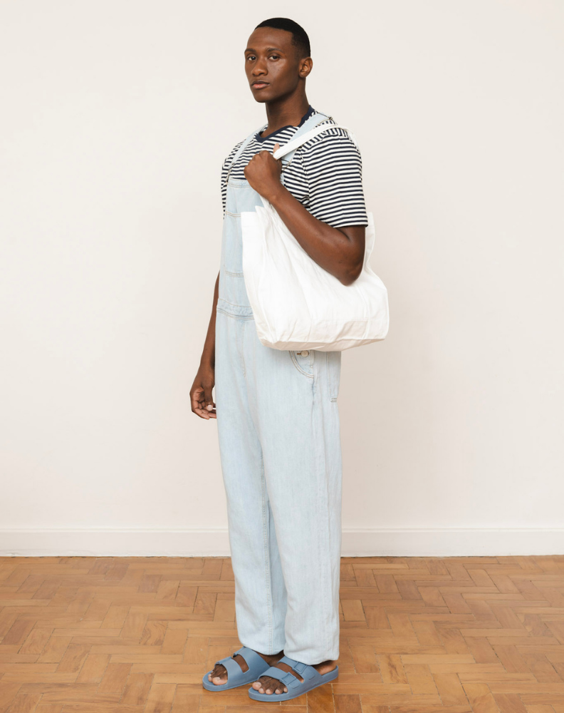 Homem de pé, usando um macacão jeans claro com uma camiseta listrada por baixo. Ele carrega uma bolsa branca e está calçando as mesmas sandálias azuis