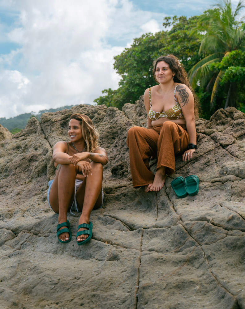 Duas mulheres sentadas em rochas em um ambiente natural. A mulher à esquerda usa um biquíni e sandálias verdes de tiras duplas, enquanto a mulher à direita veste calças marrons e um top estampado, com suas sandálias verdes colocadas ao lado.