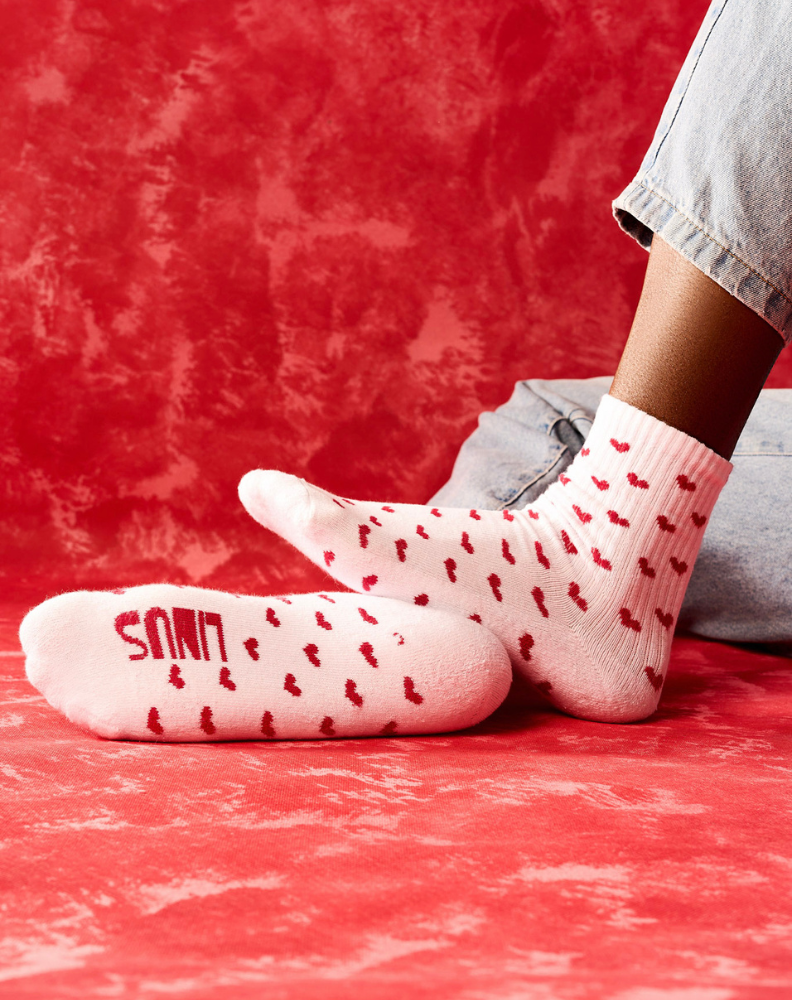 Pessoa em posição sentada com os pés usando meias de algodão estampada de corações branco e vermelho, fundo vermelho e meia com toque macio.