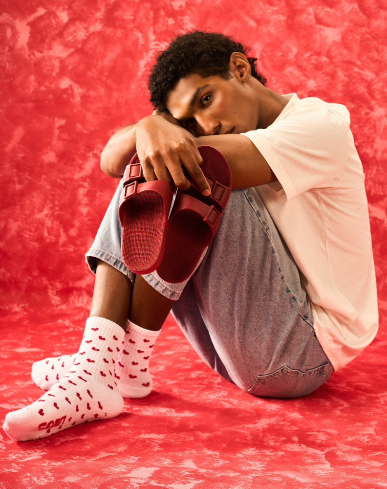 Pessoa sentada com os pés usando meias de algodão estampada de corações branco e vermelho, fundo vermelho e meia com toque macio. Nas mãos o modelo segura um par de sandálias linus bordô.