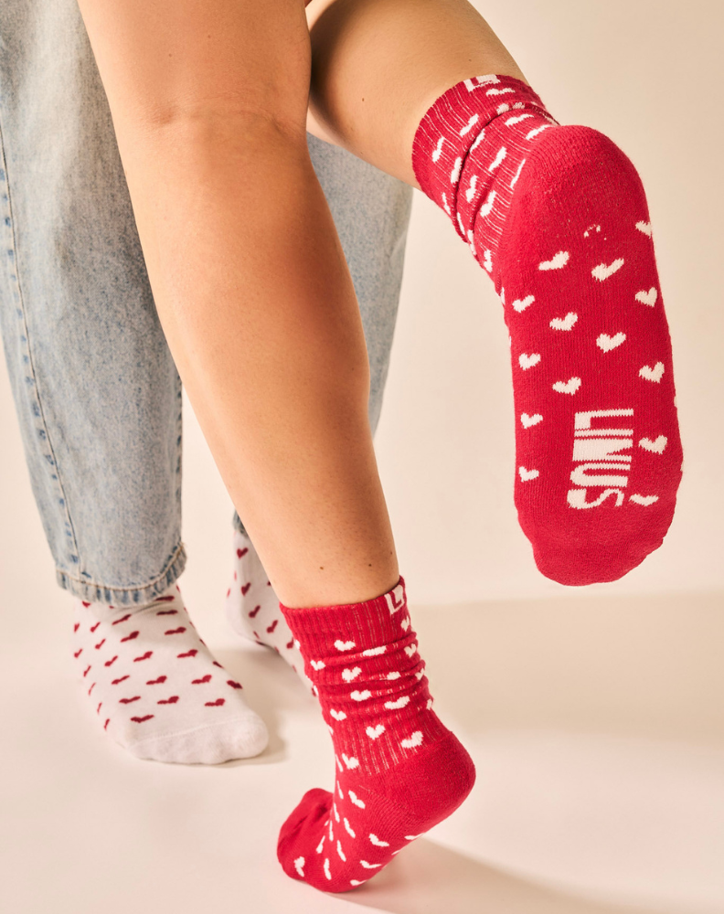 Dupla de pessoas com os pés usando meias de algodão estampada de corações vermelha e branca, fundo cinza claro e meia com toque macio.