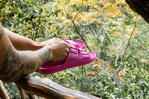 Duas mãos segurando um par de sandália rosa fúcsia apoiado em um corrimão de madeira com paisagem de floresta.