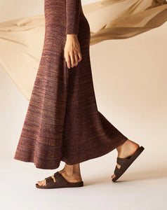 Tronco até os pés de uma mulher usando vestido em tons de marrom com um lenço beje ao fundo e com sandália marrom café