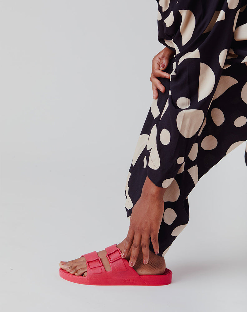 Sandália Linus Urucum com duas tiras, feita de material 100% reciclável, em pé com calça preta de estampa de bolinhas brancas