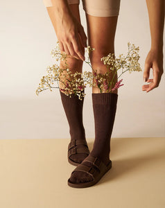 Parte inferior de uma pessoa usando meias e sandália na cor marrom café com flores dentro das meias.