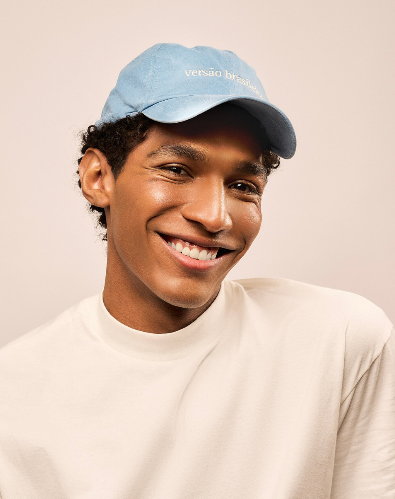 Homem sorrindo usando o boné de algodão na cor azul claro na cabeça. Bordado do boné em off white escrito Versão Brasileira.