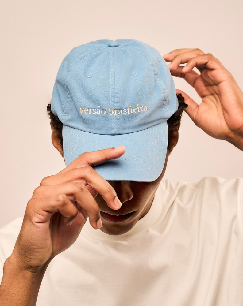Homem usando o boné de algodão na cor azul claro na cabeça e segurando a aba. Bordado do boné em off white escrito Versão Brasileira.
