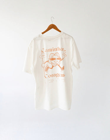 Camiseta branca pendurada em cabide com a estampa de uma flor caminhando e a frase "caminhar com coragem" com fundo branco.