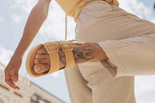 Parte inferior do corpo d euma pessoa usando calça beje e sandália amarela com um dos pés flexionado para trás.