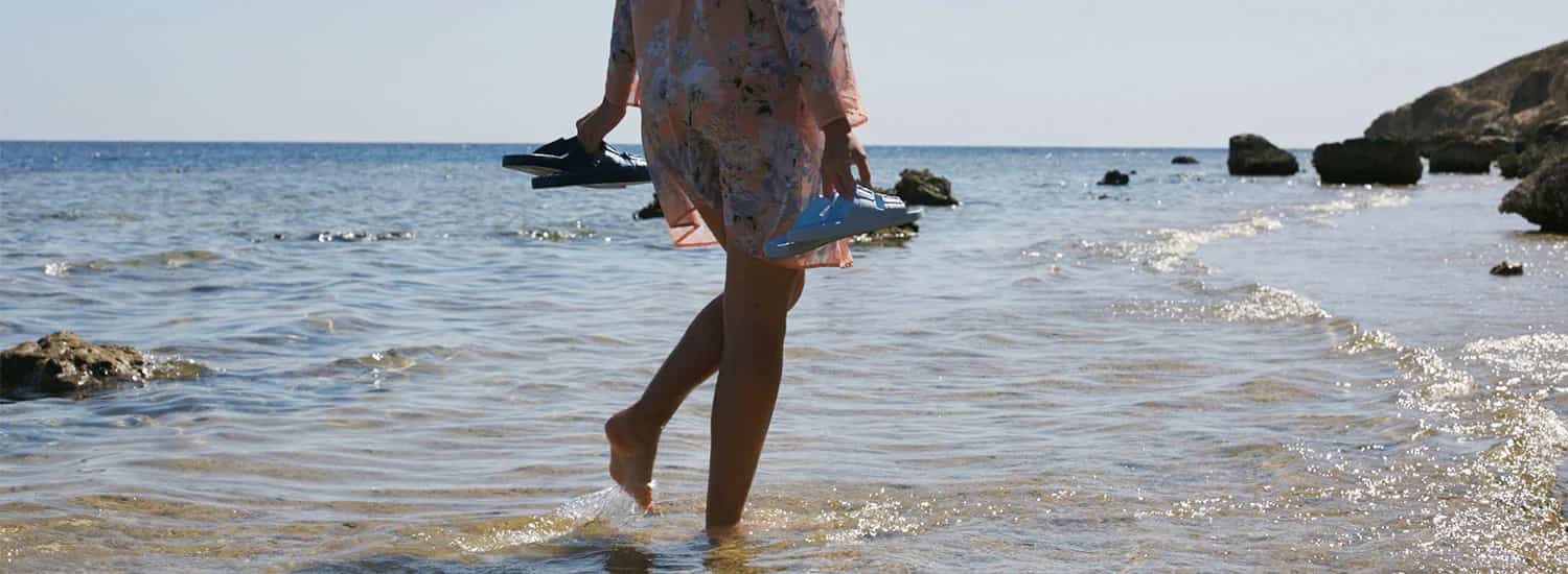 Mulher pisando no mar segurando em cada mão um par de sandália, um azul e um preto.