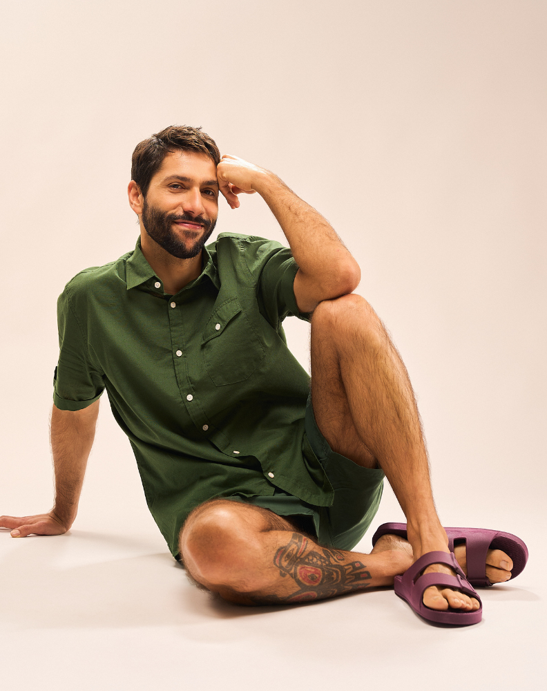Homem sentado no chão vestindo conjunto de bermuda e camisa de botão em verde combinando com as sandálias linus na cor roxo açaí. 