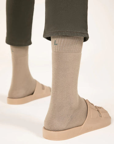 Parte inferior de duas pernas vistas de trás com calça verde usando meias e sandália na cor beje da marca Linus.