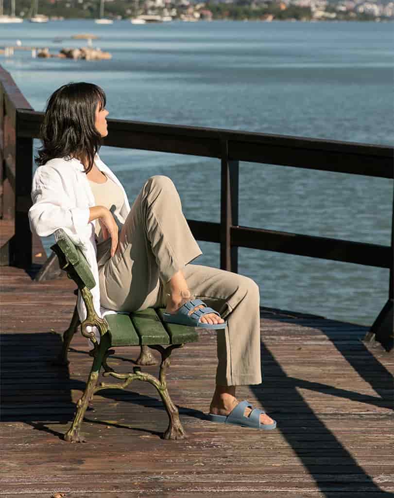 Mulher usando caça beje, blusa branca e sandália azul brisa sentada em banco verde olhando para paisagem.