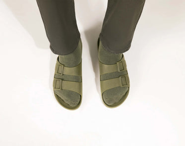Parte inferior de duas pernas vistas de frente com calça verde usando meias e sandália na cor verde musgo da marca Linus.