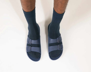 Parte inferior de duas pernas vistas de frente usando meias e sandália na cor azul da marca Linus.
