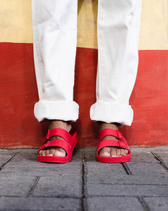 Pessoa com calça branca usando sandália na cor vermelha encontada em uma parede vermelha e amarela.