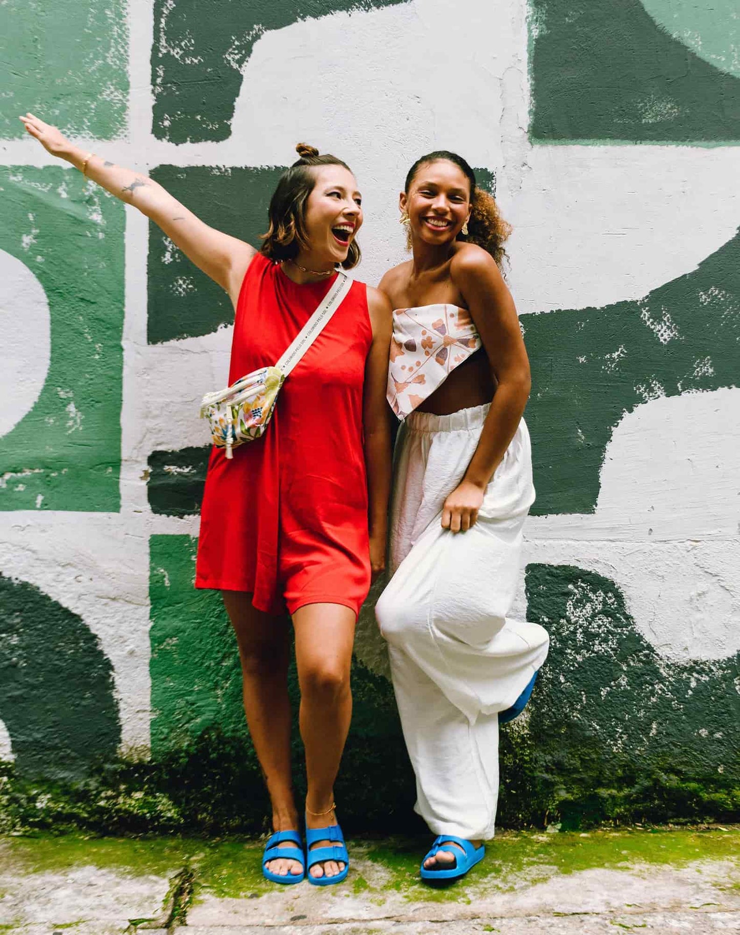 Duas mulheres abraçadas e sorrindo em frente a um muro e ambas calçando sandália azul lagoa.