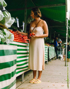 Mulher escolhendo tomates na feira, em uma barraca listrada em branco e verde, usando vestido branco e sandália vegana amarela.