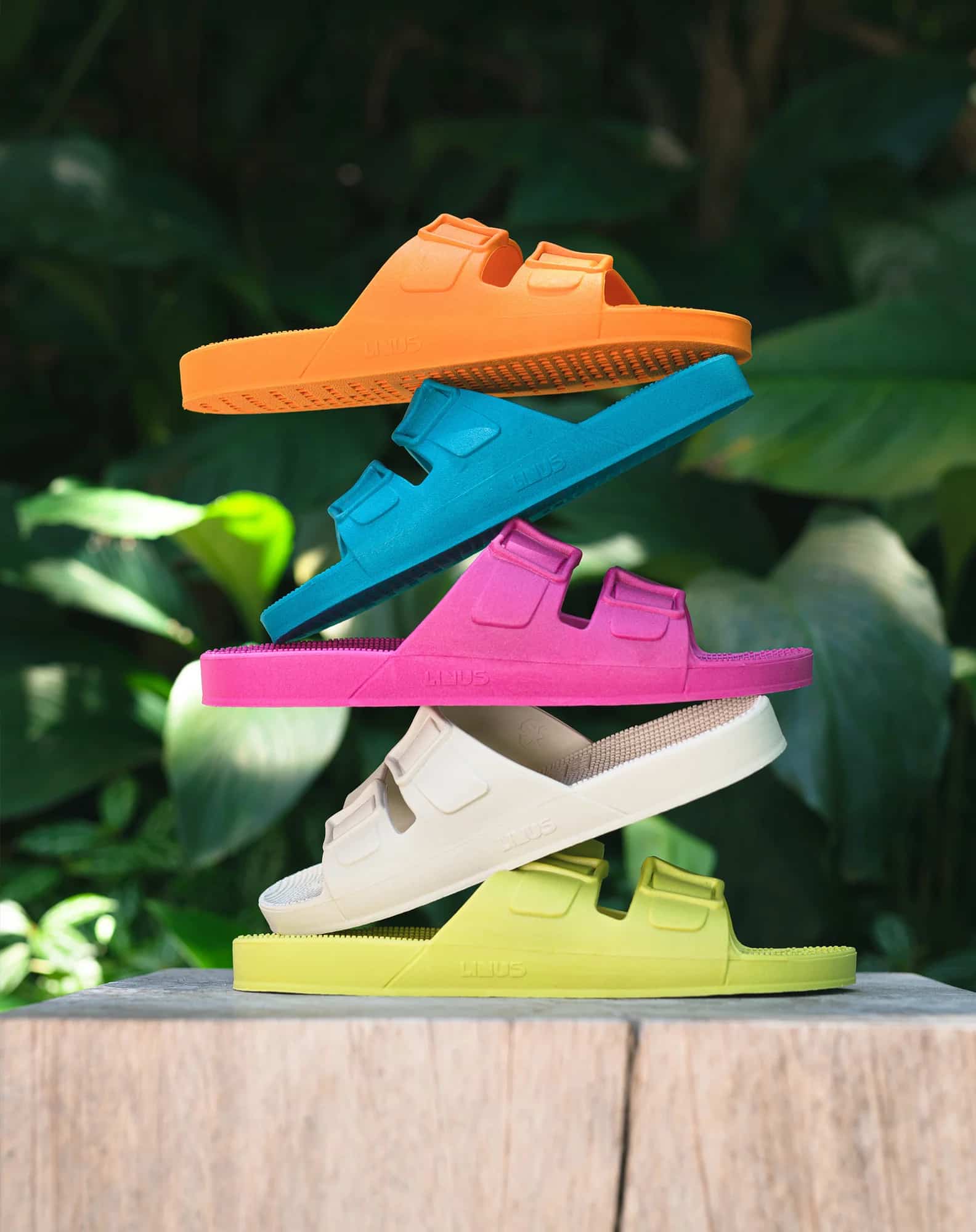 Fundo de vegetação com 5 sandálias empilhadas, de baixo para cima, verde, branca, rosa, azul e laranja.